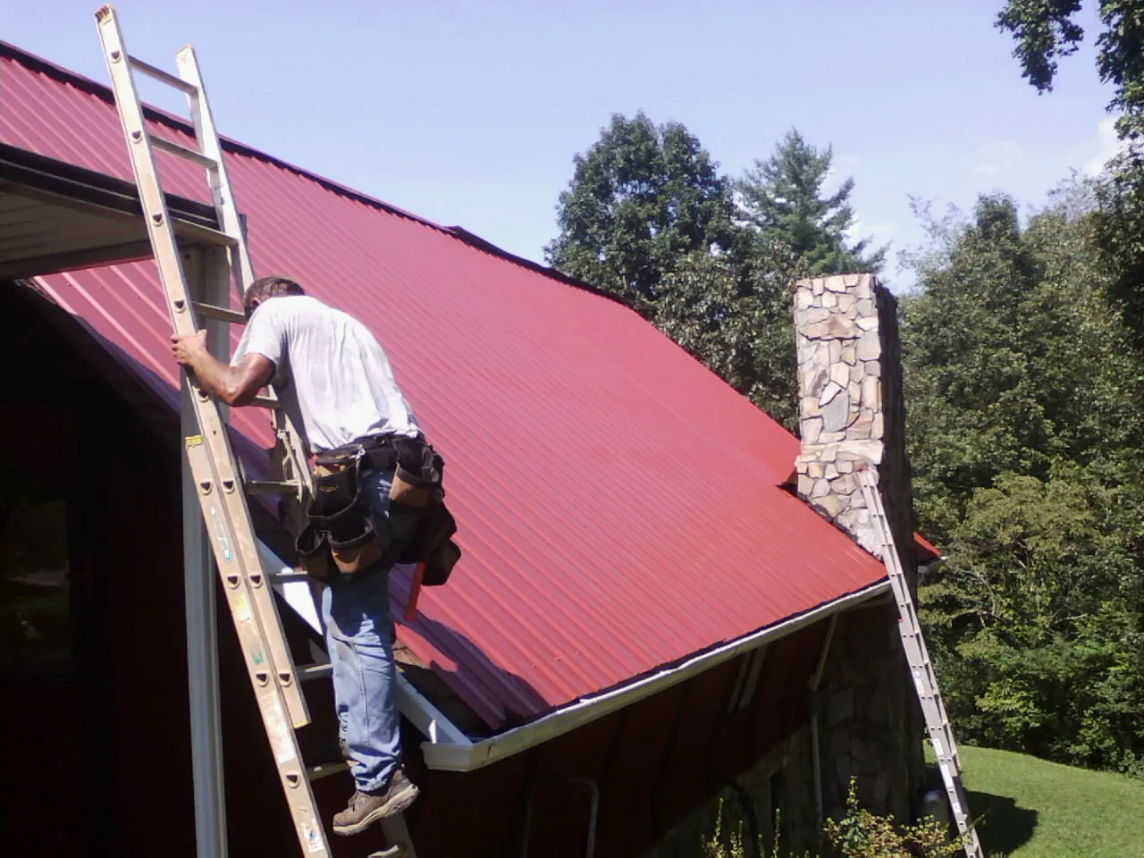Roofer on a ladder beside a red metal roof and stone chimney — Soffit & Fascia Repair by Roofing Brothers in Kennedale